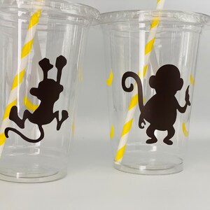 May include: Three clear plastic cups with yellow and white striped straws. Each cup has a black silhouette of a monkey with a banana on a yellow background.