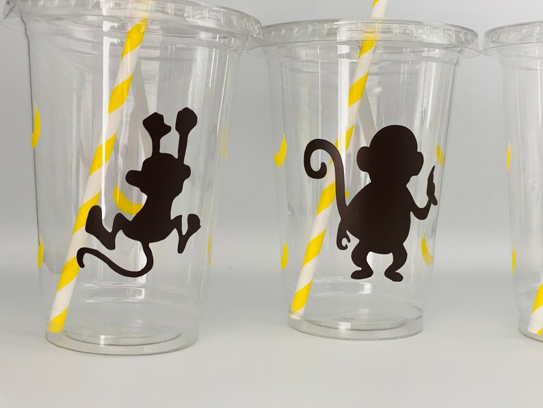 May include: Three clear plastic cups with yellow and white striped straws. Each cup has a black silhouette of a monkey with a banana on a yellow background.