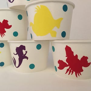 May include: Set of six white paper cups with colourful cartoon characters. The cups feature a mermaid, a yellow fish, and a red lobster. The cups are decorated with teal polka dots.