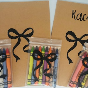 May include: Three brown paper notebooks with black ribbon bows and the name "Kacie" written on one of the notebooks. Each notebook is paired with a clear plastic bag containing a set of eight Crayola crayons.