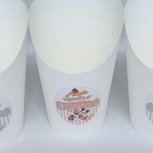 Op de afbeelding: Drie witte papieren voedselcontainers met een open bovenkant en een taps toelopende basis. Elke container is voorzien van een ronde sticker met de tekst Main Street CHURRO en een illustratie van twee tekenfilmmuizen en twee churros.