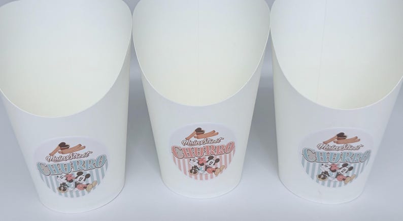 Op de afbeelding: Drie witte papieren voedselcontainers met een open bovenkant en een taps toelopende basis. Elke container is voorzien van een ronde sticker met de tekst Main Street CHURRO en een illustratie van twee tekenfilmmuizen en twee churros.