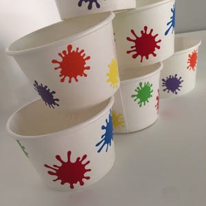 K&ouml;nnte beinhalten: Ein Stapel aus sechs wei&szlig;en Pappbechern mit bunten Farbklecksen. Die Becher sind pyramidenf&ouml;rmig gestapelt, wobei der gr&ouml;&szlig;te Becher unten und der kleinste oben liegt. Die Farbkleckse sind in verschiedenen Farben, darunter Rot, Orange, Gelb, Gr&uuml;n, Blau und Lila.