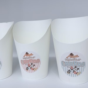 Op de afbeelding: Witte papieren voedselbakjes met een schuine bovenrand. Elk bakje is voorzien van een ronde sticker met een afbeelding van churros en stripfiguurmuizen, samen met de tekst Main Street CHURRO in een vintage lettertype op een gestreepte achtergrond.