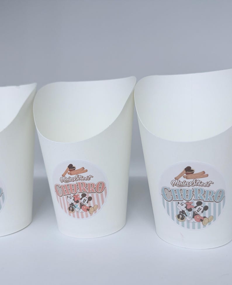 Op de afbeelding: Witte papieren voedselbakjes met een schuine bovenrand. Elk bakje is voorzien van een ronde sticker met een afbeelding van churros en stripfiguurmuizen, samen met de tekst Main Street CHURRO in een vintage lettertype op een gestreepte achtergrond.