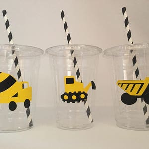 May include: Three clear plastic cups with black and white striped straws. Each cup has a different yellow and black construction vehicle design: a cement mixer, a bulldozer, and a dump truck.