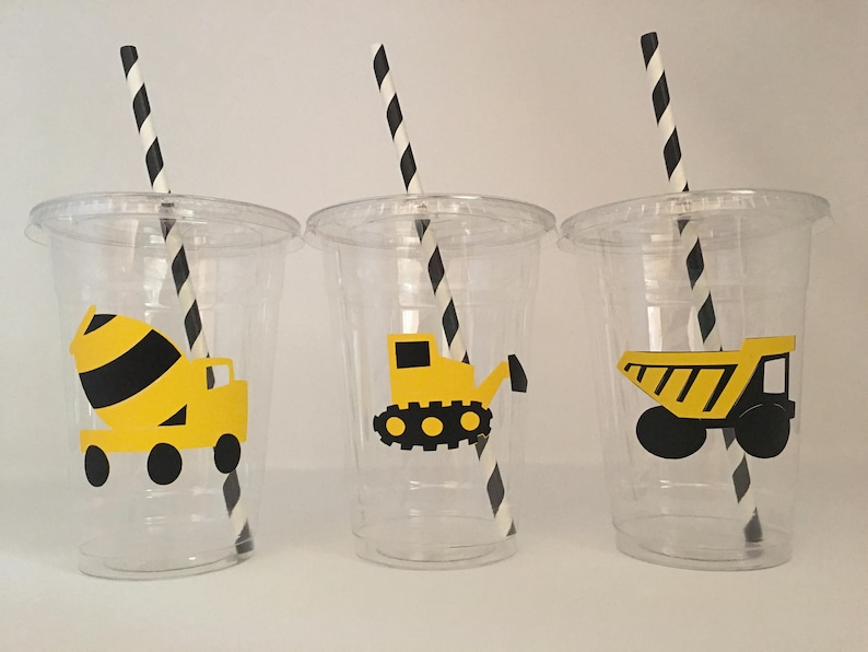 May include: Three clear plastic cups with black and white striped straws. Each cup has a different yellow and black construction vehicle design: a cement mixer, a bulldozer, and a dump truck.