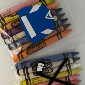 May include: A clear plastic bag filled with a variety of Crayola crayons. The crayons are arranged in a row, with the tips facing up. There is a blue hockey jersey and a black hockey stick with white tape on the top of the crayons.