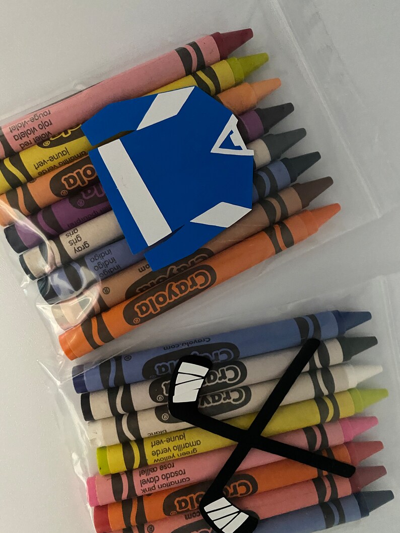 May include: A clear plastic bag filled with a variety of Crayola crayons. The crayons are arranged in a row, with the tips facing up. There is a blue hockey jersey and a black hockey stick with white tape on the top of the crayons.