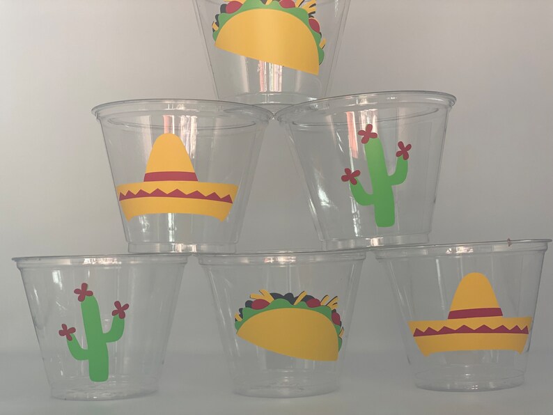 May include: Six clear plastic cups with different Mexican-themed designs. The designs include a sombrero, a cactus, and a taco.