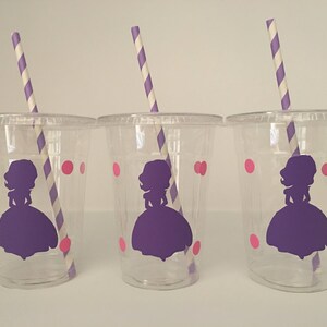 May include: Three clear plastic cups with purple and pink designs. Each cup has a purple silhouette of a princess with a crown and a pink polka dot pattern. Each cup has a striped straw.