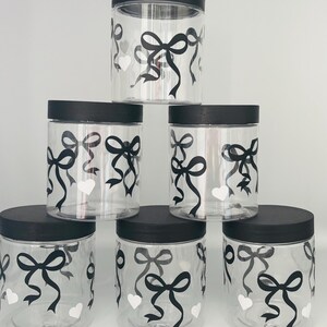 May include: Six clear plastic jars with black lids are stacked. Each jar is decorated with black bows and white hearts. The jars are cylindrical and appear to be empty.