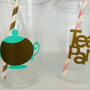 Puede incluir: Vasos de pl&aacute;stico transparente con elementos decorativos para una fiesta de t&eacute;. Un vaso presenta una tetera marr&oacute;n con tapa y boquilla turquesa, y una pajita a rayas marrones y blancas. El otro vaso tiene las palabras "Tea Party" en dorado.