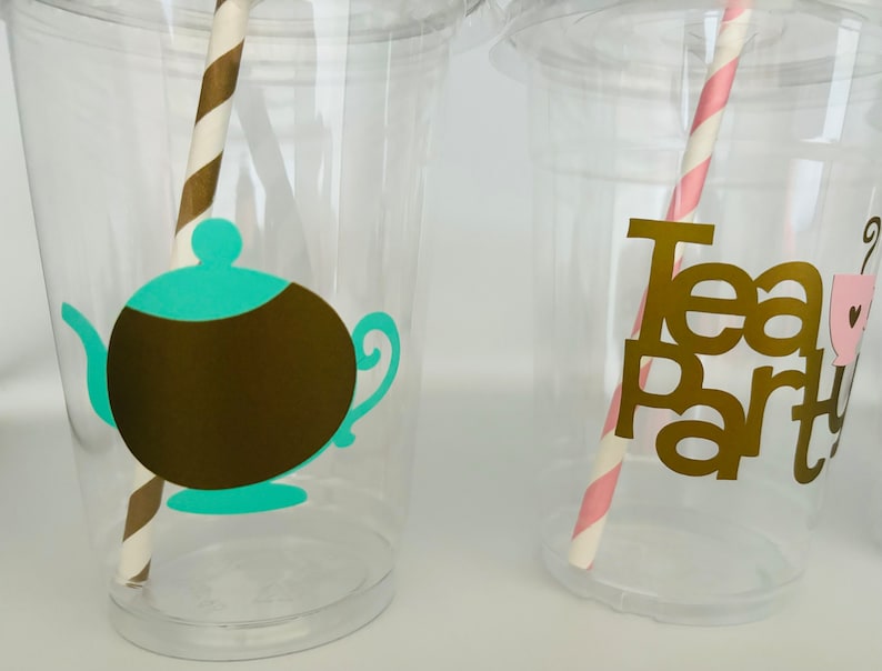Puede incluir: Vasos de pl&aacute;stico transparente con elementos decorativos para una fiesta de t&eacute;. Un vaso presenta una tetera marr&oacute;n con tapa y boquilla turquesa, y una pajita a rayas marrones y blancas. El otro vaso tiene las palabras "Tea Party" en dorado.