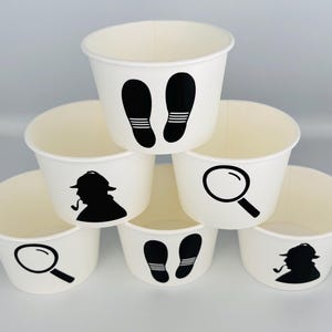 Puede incluir: Seis vasos de papel blancos con siluetas negras de una lupa, huellas y un detective. Los vasos est&aacute;n apilados en forma de pir&aacute;mide.