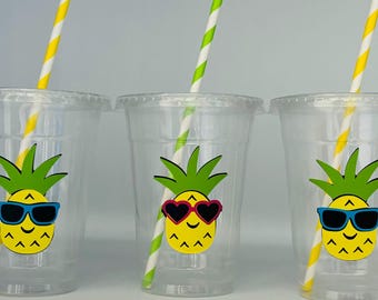 May include: Three clear plastic cups with yellow and white striped straws. Each cup has a pineapple with sunglasses and a smiling face. The pineapple in the middle cup has pink heart-shaped sunglasses.