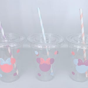 May include: Three clear plastic cups with lids and straws. Each cup features a Minnie Mouse design with a bow and polka dots in pastel colors. The straws have striped patterns in various colors.