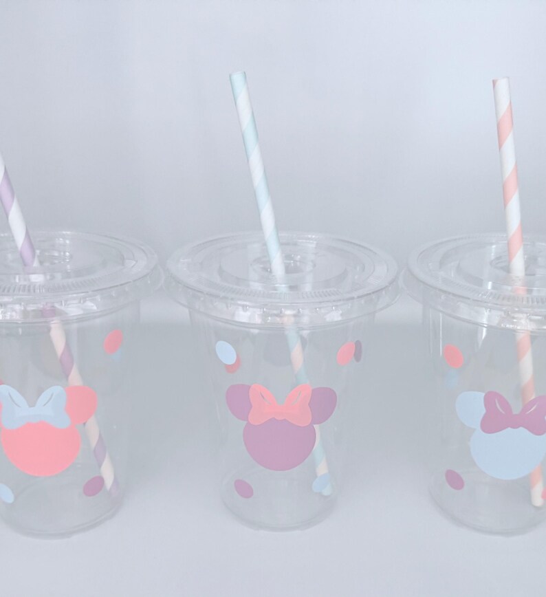 May include: Three clear plastic cups with lids and straws. Each cup features a Minnie Mouse design with a bow and polka dots in pastel colors. The straws have striped patterns in various colors.