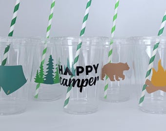 May include: Clear plastic cups with green and white striped straws. Each cup features a camping-themed graphic, including a tent, trees, a bear, a campfire, and the words "Happy Camper" in black.