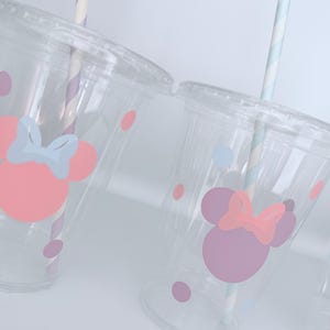 May include: Clear plastic cups with lids, decorated with pink and purple Minnie Mouse designs and polka dots. Each cup has a striped paper straw. The cups are designed for cold beverages.