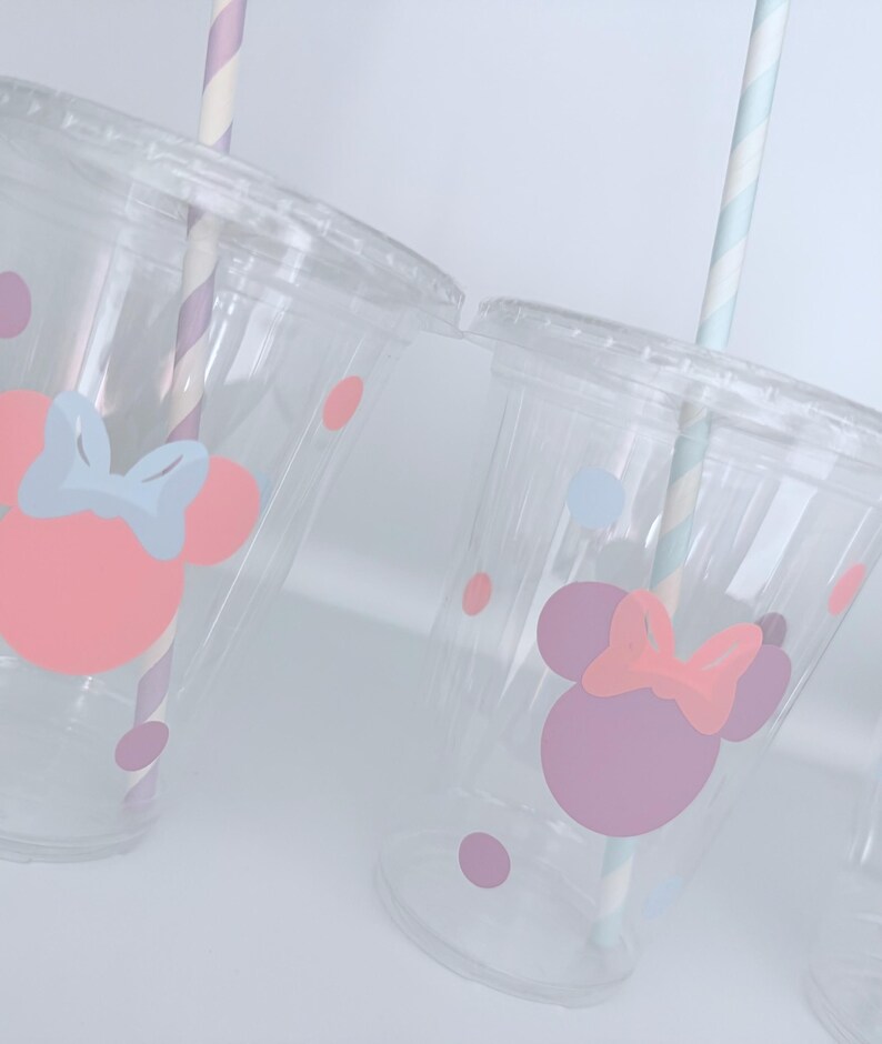 May include: Clear plastic cups with lids, decorated with pink and purple Minnie Mouse designs and polka dots. Each cup has a striped paper straw. The cups are designed for cold beverages.