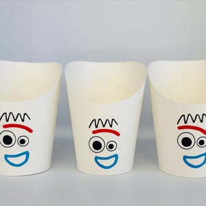 May include: Three off-white paper cups with a cartoon face design. Each cup features a black squiggly hair design, a red eyebrow, two black eyes, and a blue smile. The cups are arranged side by side.