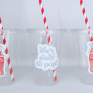 May include: Three clear plastic cups with red and white striped straws. Each cup has a sticker with a Coca-Cola bottle design. One cup has the text "she's ready to pop!"