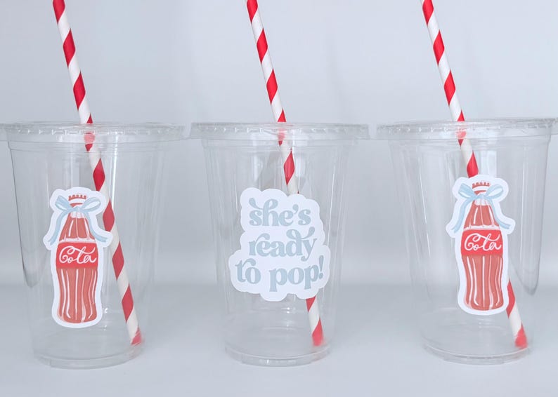 May include: Three clear plastic cups with red and white striped straws. Each cup has a sticker with a Coca-Cola bottle design. One cup has the text "she's ready to pop!"