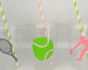Op de afbeelding: Drie doorzichtige plastic bekers met rietjes en tennis-thema ontwerpen. Eén beker heeft een tennisracket, één heeft een tennisbal en één heeft een tennisspeler silhouet.