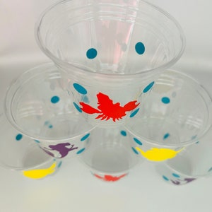 Puede incluir: Pila de vasos de pl&aacute;stico transparente con lunares azules y siluetas de animales de colores. El vaso de arriba tiene una langosta roja, el siguiente vaso tiene un dinosaurio morado, el siguiente vaso tiene un p&aacute;jaro amarillo y el vaso de abajo tiene un dinosaurio morado.