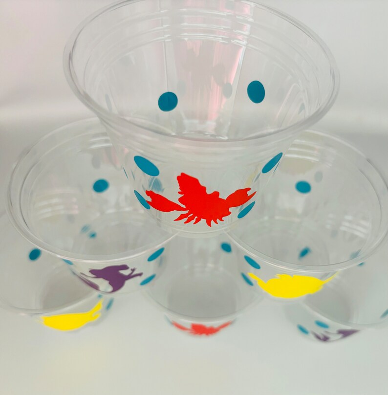 Puede incluir: Pila de vasos de pl&aacute;stico transparente con lunares azules y siluetas de animales de colores. El vaso de arriba tiene una langosta roja, el siguiente vaso tiene un dinosaurio morado, el siguiente vaso tiene un p&aacute;jaro amarillo y el vaso de abajo tiene un dinosaurio morado.