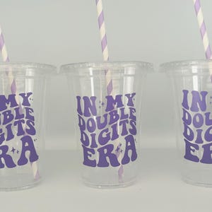 May include: Three clear plastic cups with purple text that reads "In My Double Digits Era" and a striped straw in each cup.