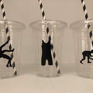 K&ouml;nnte beinhalten: Drei durchsichtige Plastikbecher mit schwarz-wei&szlig; gestreiften Strohhalmen. Jeder Becher hat eine schwarze Silhouette einer anderen Wrestling-Pose.