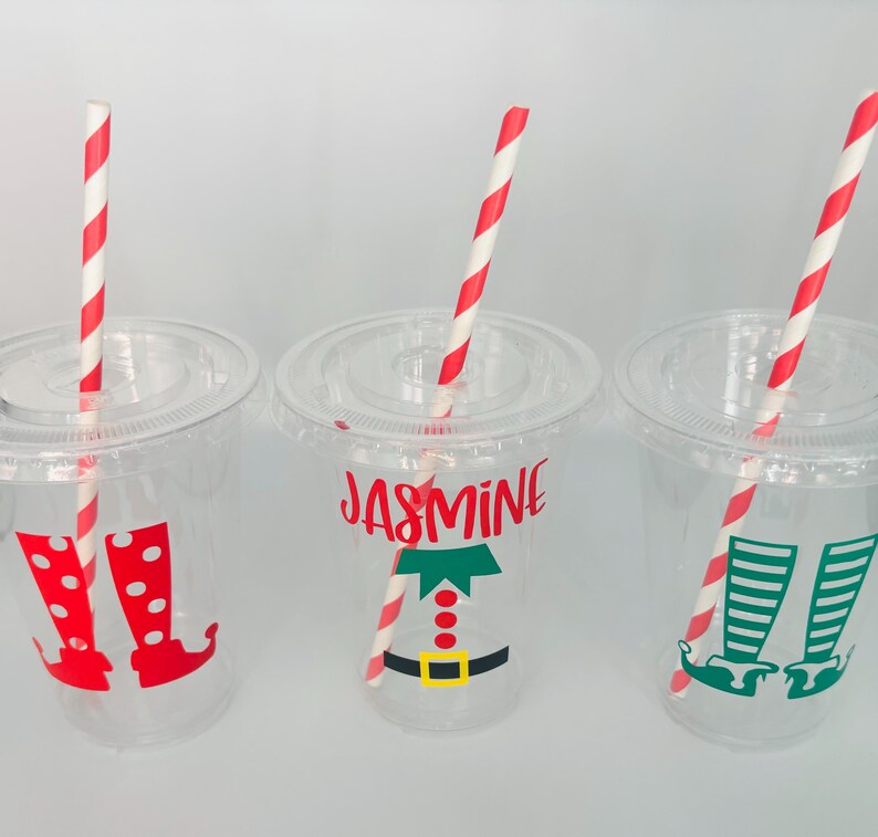 May include: Three clear plastic cups with red and white striped straws. Each cup has a different elf design with the name "Jasmine" on one cup.