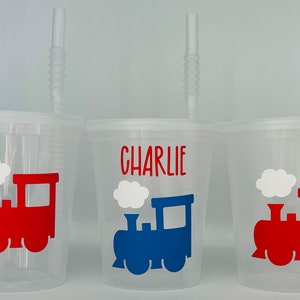 May include: Three clear plastic cups with straws and a train design. The cups have a red train, a blue train, and a red train. The middle cup has the name "CHARLIE" printed on it.