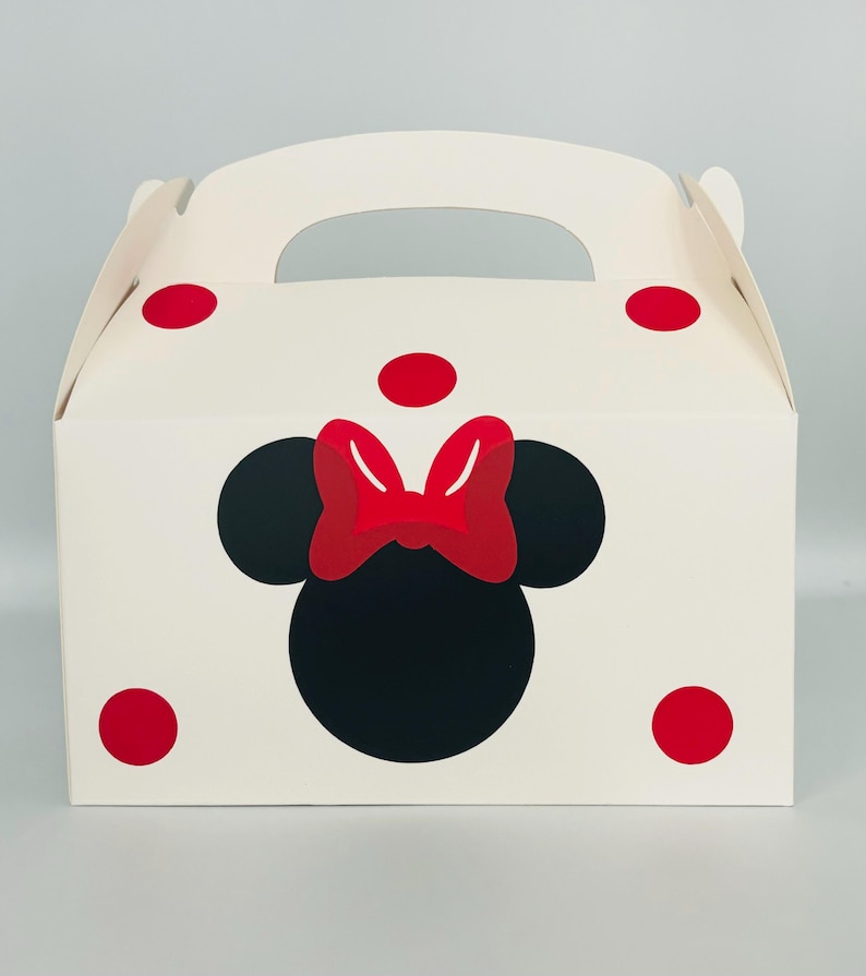 Puede incluir: Caja de regalo de cart&oacute;n blanco con un patr&oacute;n de lunares rojos. La caja tiene un asa y presenta una silueta de Minnie Mouse negra y roja.
