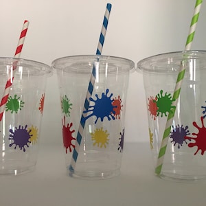 May include: Three clear plastic cups with colorful paint splatter designs and matching striped straws. The straws are red, blue, and green.