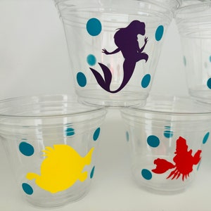 Puede incluir: Vasos de pl&aacute;stico transparentes con lunares color turquesa y varios personajes de Disney, incluyendo una sirena, un cangrejo y un pez.