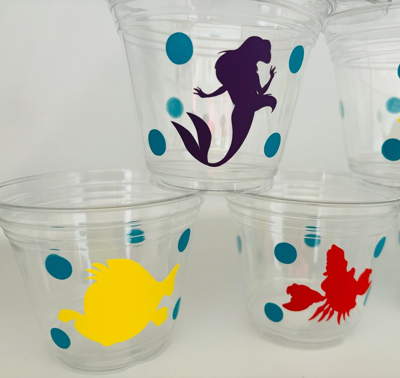 Puede incluir: Vasos de pl&aacute;stico transparentes con lunares color turquesa y varios personajes de Disney, incluyendo una sirena, un cangrejo y un pez.