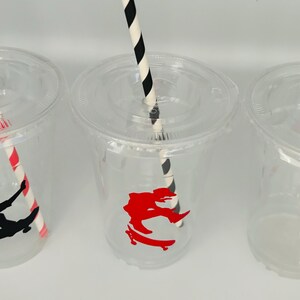 Puede incluir: Tres vasos de pl&aacute;stico transparentes con pajitas rayadas rojas y negras. Cada vaso tiene una pegatina con la silueta de una persona haciendo skate. Una pegatina es roja y las otras dos son negras.