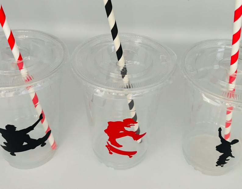 Puede incluir: Tres vasos de pl&aacute;stico transparentes con pajitas rayadas rojas y negras. Cada vaso tiene una pegatina con la silueta de una persona haciendo skate. Una pegatina es roja y las otras dos son negras.