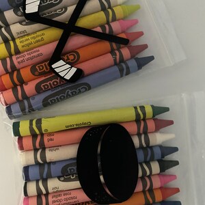 May include: A clear plastic bag filled with a set of Crayola crayons. The crayons are arranged in a row, with the tips facing the viewer. The bag has a hockey stick and puck design printed on it.