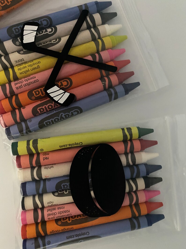 May include: A clear plastic bag filled with a set of Crayola crayons. The crayons are arranged in a row, with the tips facing the viewer. The bag has a hockey stick and puck design printed on it.