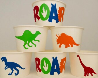 May include: White paper cups with dinosaur designs. Some cups have the word "ROAR" in red, green, blue, and orange. Other cups feature green, orange, blue, and red dinosaur silhouettes. Perfect for a dinosaur-themed party.