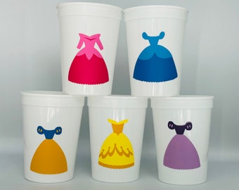 May include: Set of five white plastic cups with different princess dress designs. The dresses are pink, blue, yellow, purple, and gold.