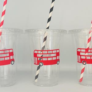 May include: Three clear plastic cups with red and white striped straws. Each cup has a red double-decker bus sticker on the front.