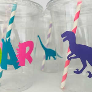 May include: Clear plastic cups with lids and striped straws. The cups are decorated with dinosaur silhouettes and the word "ROAR" in bright colors. A purple T-Rex dinosaur is featured on one cup.