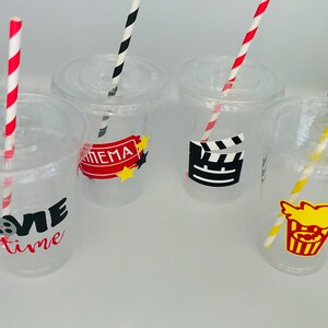 May include: Four clear plastic cups with red and white striped straws. Each cup has a different movie-themed design: "Movie Time", "Cinema", a film clapboard, and a popcorn bucket.