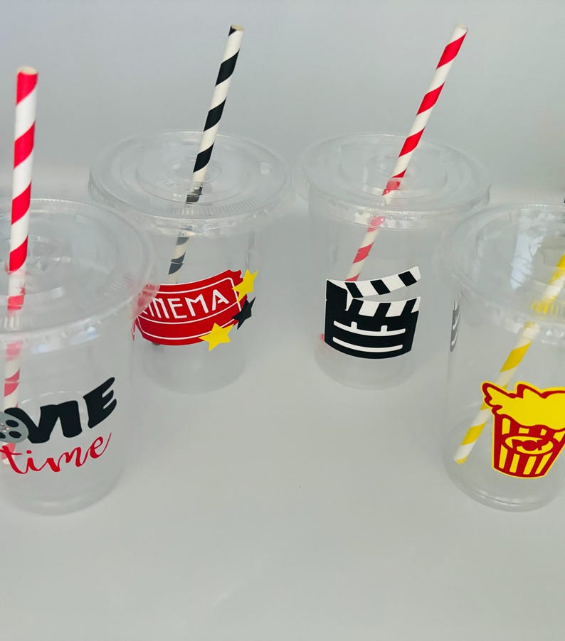 May include: Four clear plastic cups with red and white striped straws. Each cup has a different movie-themed design: "Movie Time", "Cinema", a film clapboard, and a popcorn bucket.