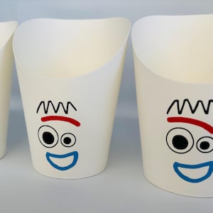 May include: Three white paper cups with a cartoon face design. Each cup features a black zig-zag hair design, a red curved line, two black eyes, and a blue smiling mouth. The cups are empty and have a curved top.
